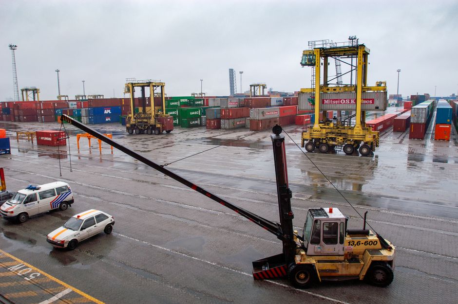 Container terminal with straddle carriers and stacked shipping containers under a cloudy sky.